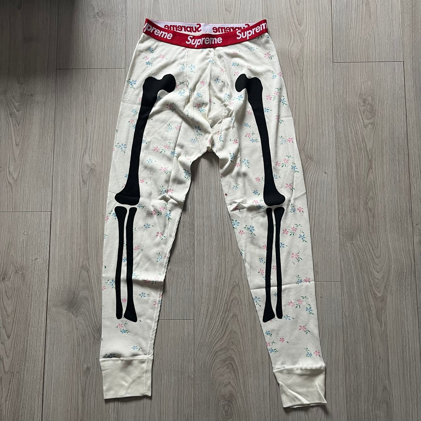 Supreme Hanes Bones Thermal Pant FW25 in natural/white with small floral all over print, crafted from 100% cotton. Features black skeletal bone prints running down both legs and a red elastic waistband with white Supreme branding throughout. Released October 23, 2025.