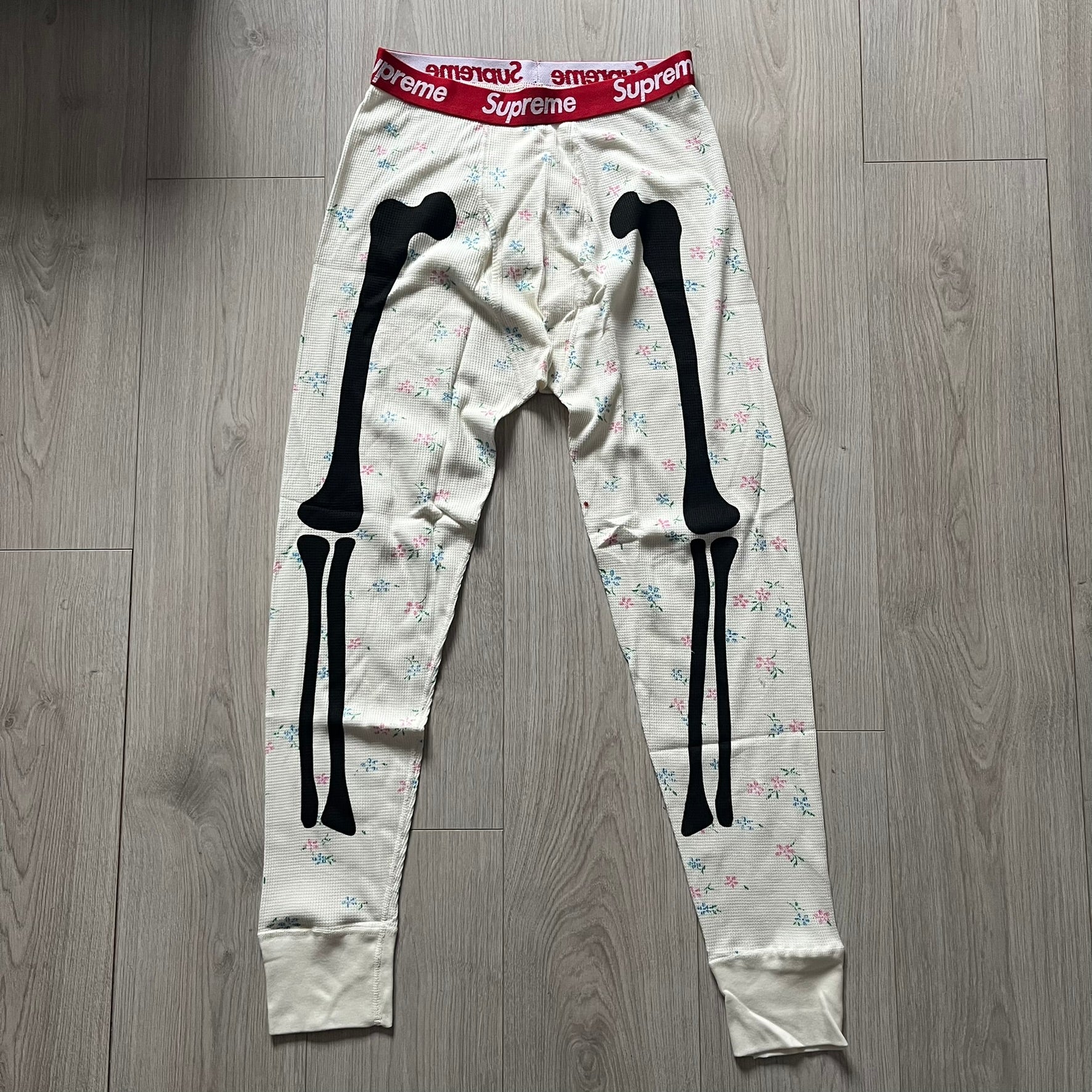 Supreme Hanes Bones Thermal Pant FW25 in natural/white with small floral all over print, crafted from 100% cotton. Features black skeletal bone prints running down both legs and a red elastic waistband with white Supreme branding throughout. Released October 23, 2025.