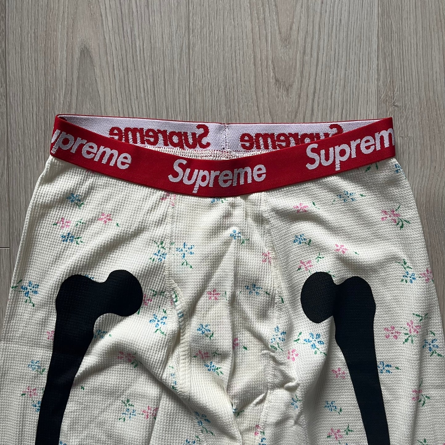 Supreme Hanes Bones Thermal Pant FW25 in natural/white with small floral all over print, crafted from 100% cotton. Features black skeletal bone prints running down both legs and a red elastic waistband with white Supreme branding throughout. Released October 23, 2025.