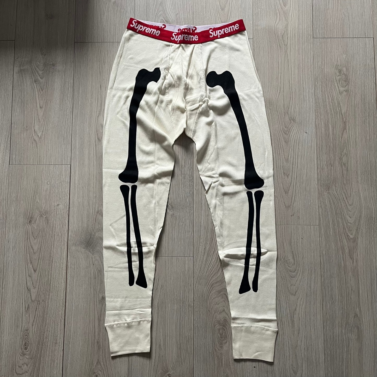 Supreme Hanes Bones Thermal Pant FW25 in natural/white, crafted from 100% cotton. Features black skeletal bone prints running down both legs and a red elastic waistband with white Supreme branding throughout. Released October 23, 2025.