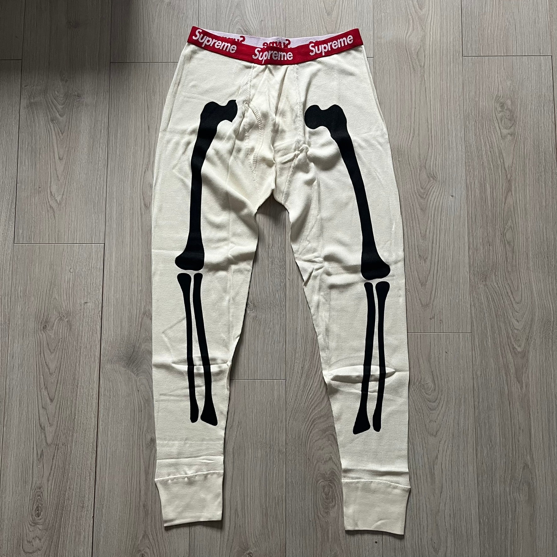 Supreme Hanes Bones Thermal Pant FW25 in natural/white, crafted from 100% cotton. Features black skeletal bone prints running down both legs and a red elastic waistband with white Supreme branding throughout. Released October 23, 2025.