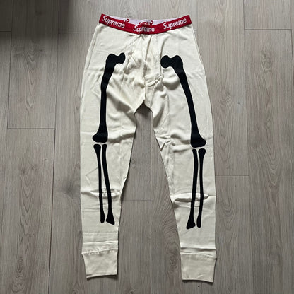Supreme Hanes Bones Thermal Pant FW25 in natural/white, crafted from 100% cotton. Features black skeletal bone prints running down both legs and a red elastic waistband with white Supreme branding throughout. Released October 23, 2025.