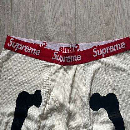 Supreme Hanes Bones Thermal Pant FW25 in natural/white, crafted from 100% cotton. Features black skeletal bone prints running down both legs and a red elastic waistband with white Supreme branding throughout. Released October 23, 2025.