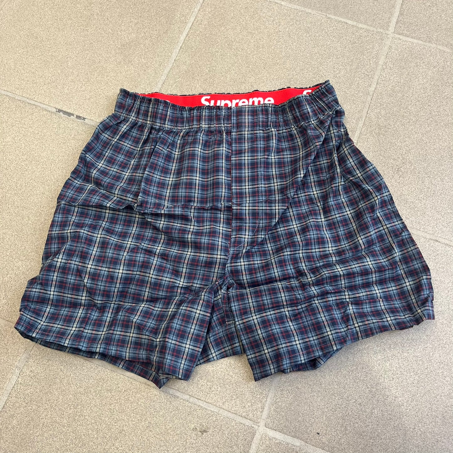 Supreme Hanes FW26 Plaid Boxers feature a plaid pattern and a red Supreme elastic waistband concealed inside. Made from 55% cotton and 45% polyester with Hanes Comfort Flex construction. Pink, grey, blue, and yellow colorways.