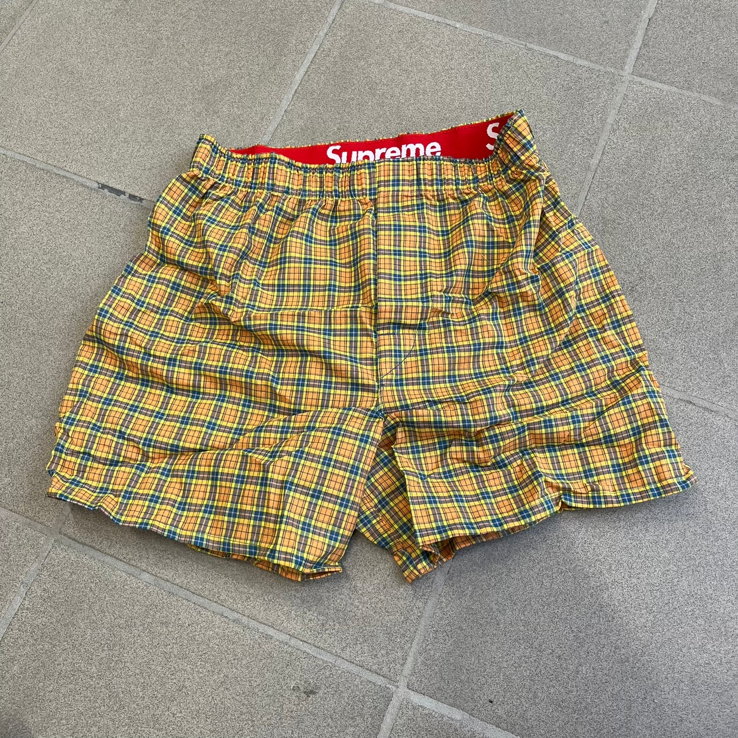 Supreme Hanes FW26 Plaid Boxers feature a plaid pattern and a red Supreme elastic waistband concealed inside. Made from 55% cotton and 45% polyester with Hanes Comfort Flex construction. Pink, grey, blue, and yellow colorways.