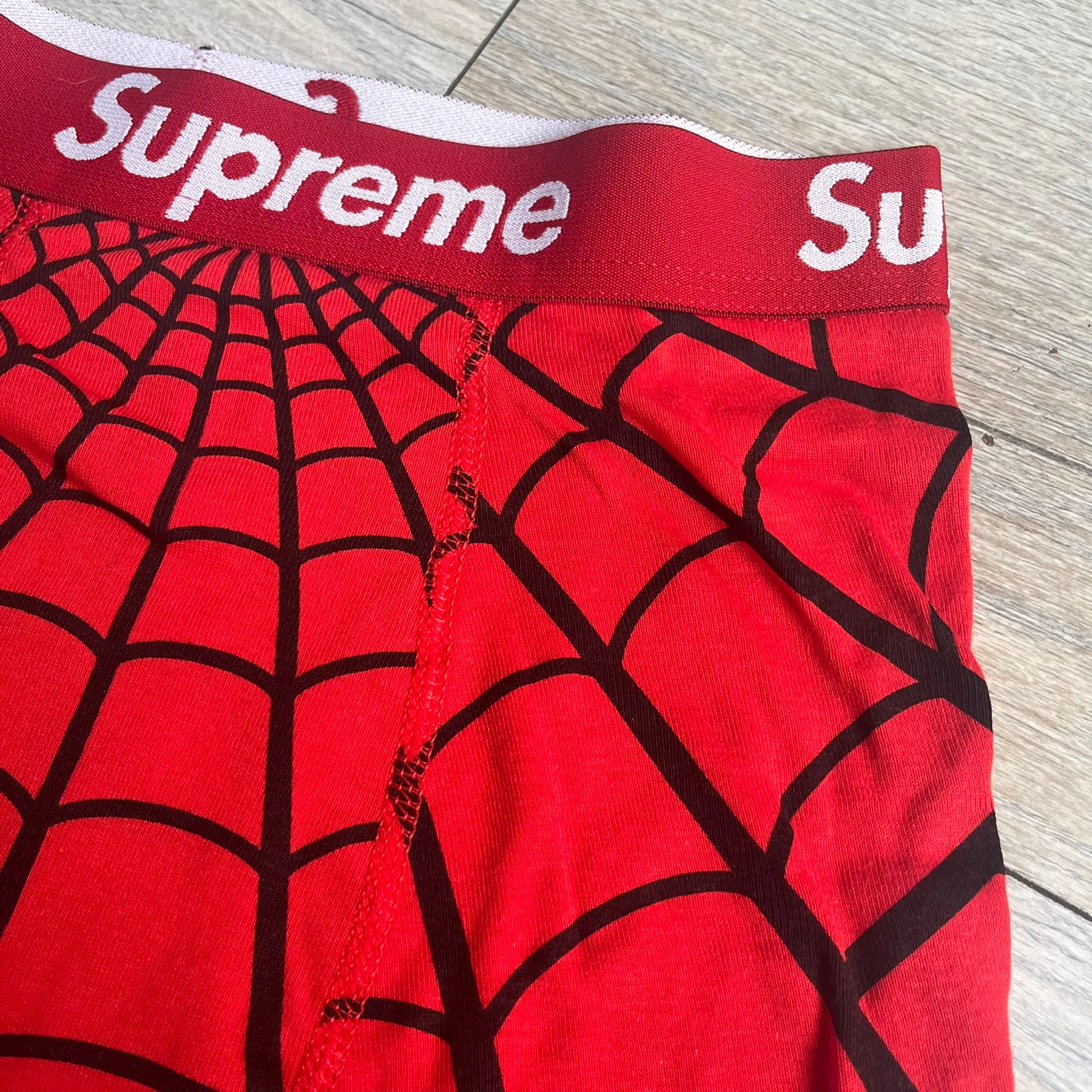 Supreme Hanes SS26 Spider-Man Red Boxer Briefs are made of cotton with a stretch fit. Features a red elastic waistband with white Supreme logo text and an all-over black web print. Classic Hanes base model a Spider-Man twist.
