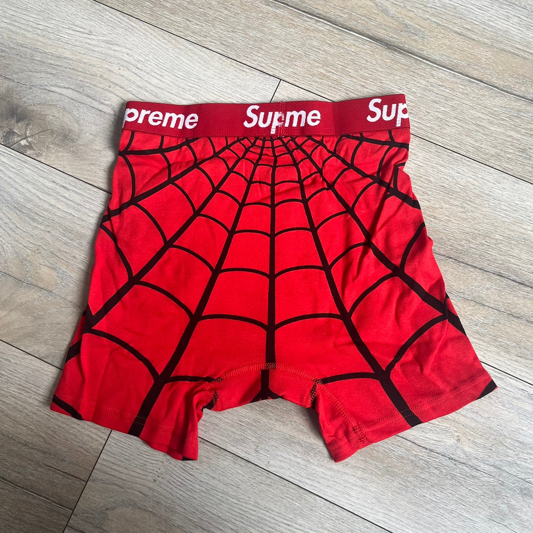 Supreme Hanes SS26 Spider-Man Red Boxer Briefs are made of cotton with a stretch fit. Features a red elastic waistband with white Supreme logo text and an all-over black web print. Classic Hanes base model a Spider-Man twist.