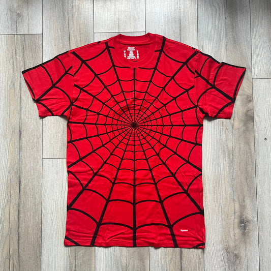 Supreme Hanes SS26 Spider-Man Red Tagless Tee is a red all-cotton crewneck T-shirt from the Spring/Summer 2026 collection, produced on a Hanes tagless blank. The shirt features an all-over black web print covering the front, back, and sleeves, with a small red Supreme stamp logo printed near the bottom corner.