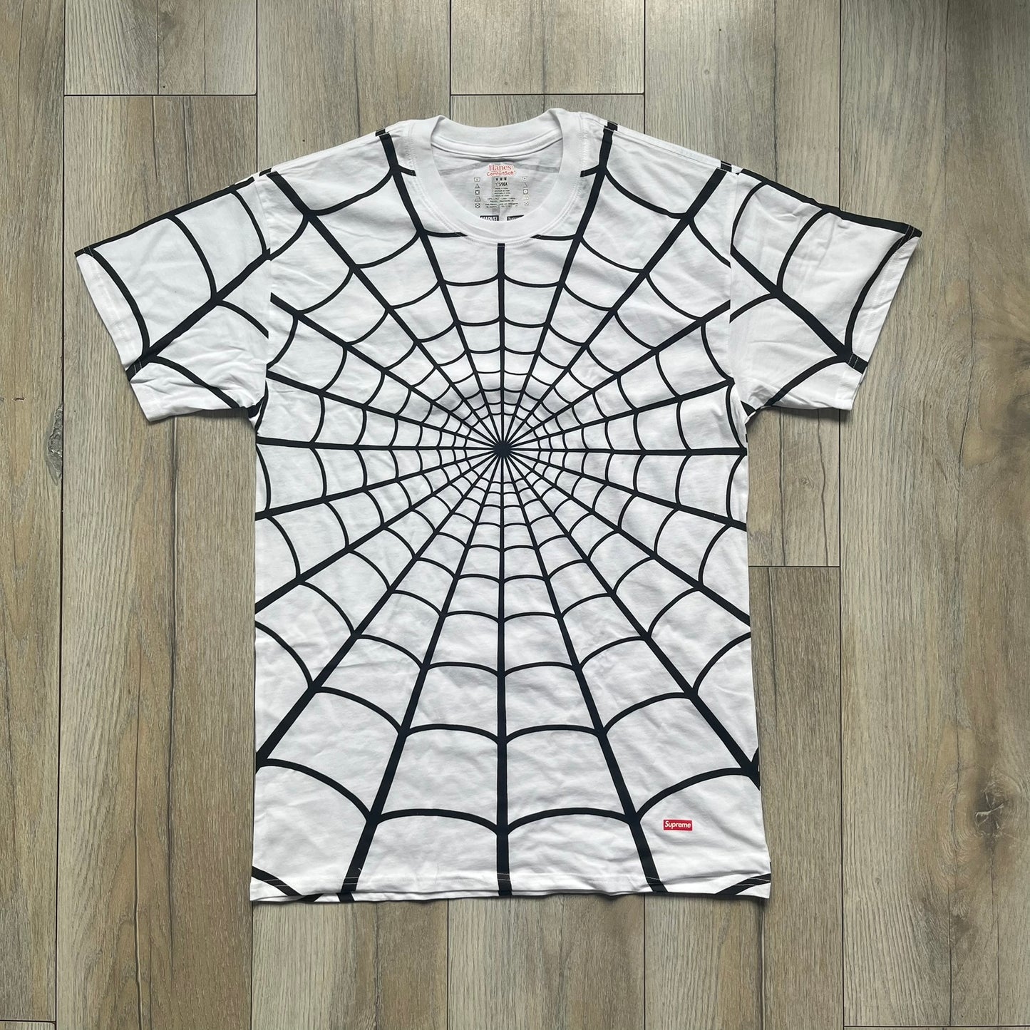 Supreme Hanes SS26 Spider-Man White Tagless Tee is a white all-cotton crewneck T-shirt from the Spring/Summer 2026 collection, produced on a Hanes tagless blank. The shirt features an all-over black web print covering the front, back, and sleeves, with a small red Supreme stamp logo printed near the bottom corner.