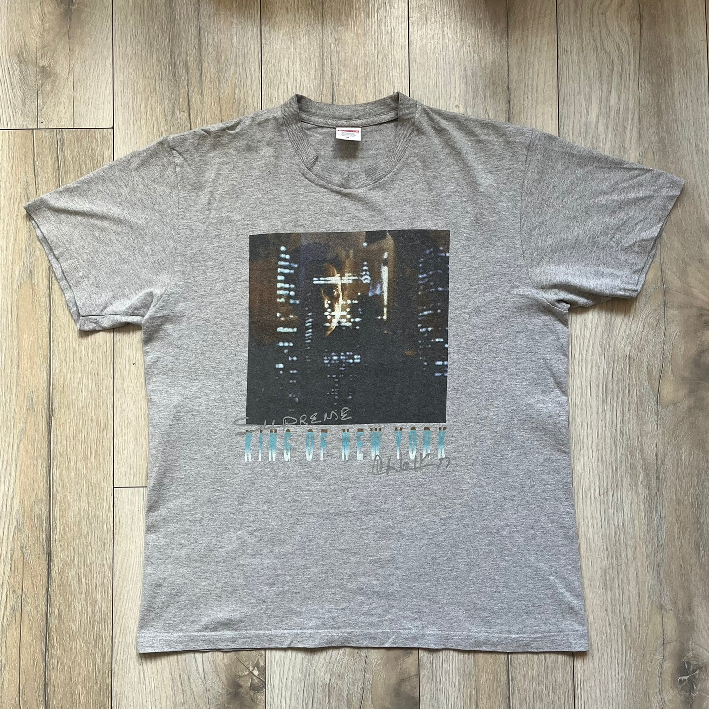 Supreme King of New York SS19 Gray Tee is a heather gray T-shirt released on February 21, 2019, as part of the SS19 collection. The front features an edited King of New York movie cover graphic with Christopher Walken’s name removed and replaced by handwritten-style “Supreme” and “C. Walken” text. The print is based on the original film cover artwork.
