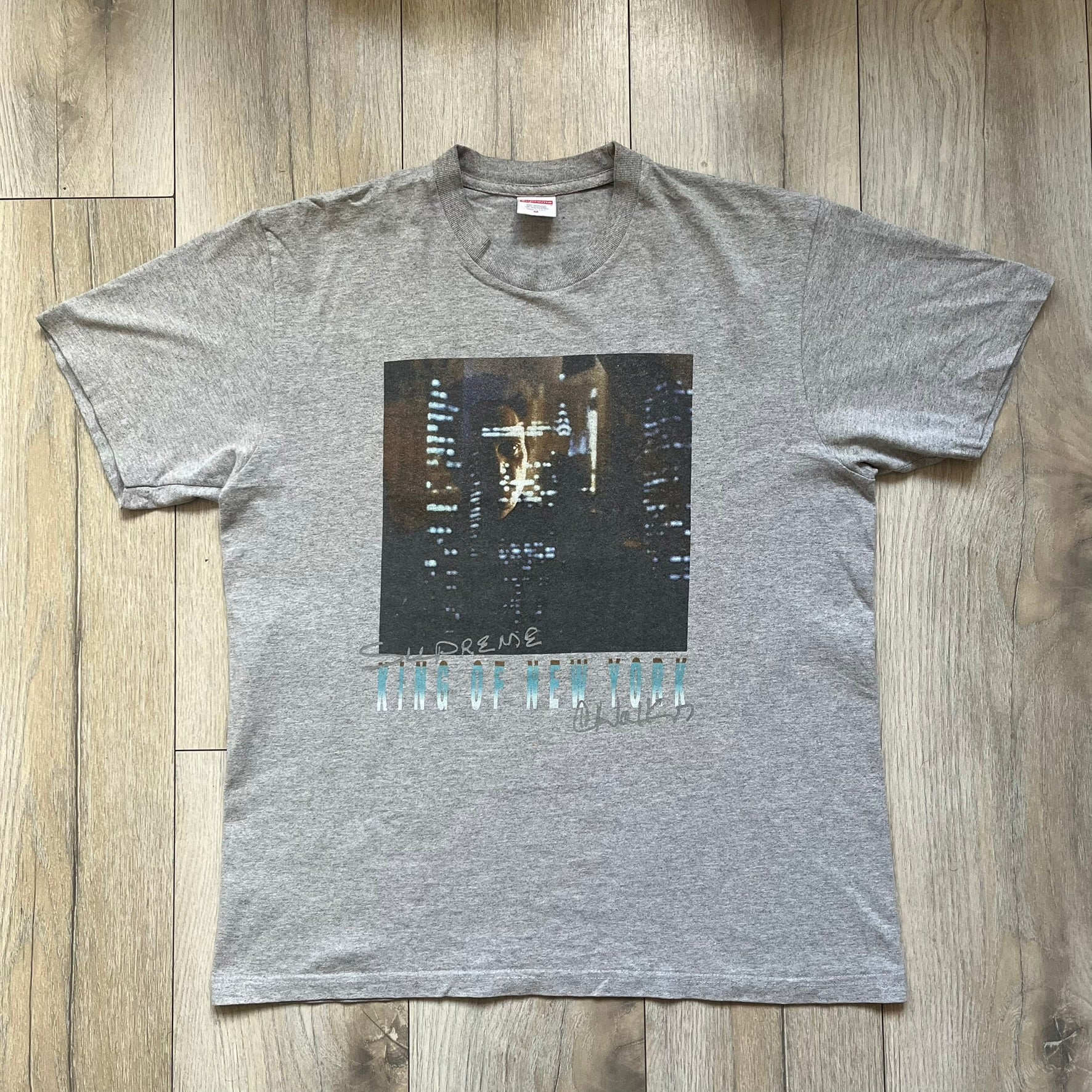 Supreme King of New York SS19 Gray Tee is a heather gray T-shirt released on February 21, 2019, as part of the SS19 collection. The front features an edited King of New York movie cover graphic with Christopher Walken’s name removed and replaced by handwritten-style “Supreme” and “C. Walken” text. The print is based on the original film cover artwork.