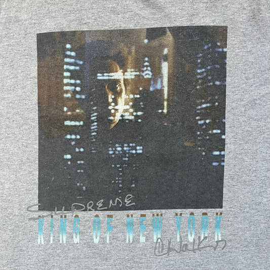 Supreme King of New York SS19 Gray Tee is a heather gray T-shirt released on February 21, 2019, as part of the SS19 collection. The front features an edited King of New York movie cover graphic with Christopher Walken’s name removed and replaced by handwritten-style “Supreme” and “C. Walken” text. The print is based on the original film cover artwork.