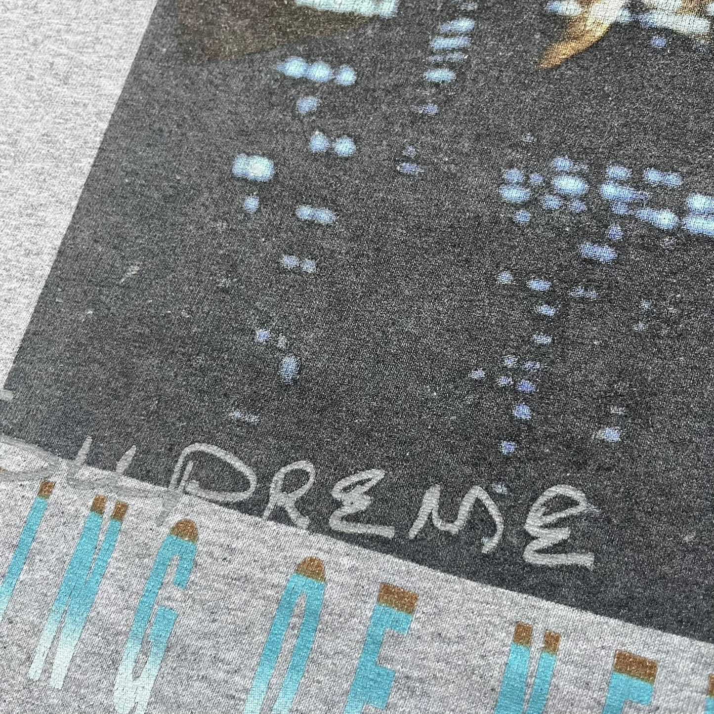 Supreme King of New York SS19 Gray Tee is a heather gray T-shirt released on February 21, 2019, as part of the SS19 collection. The front features an edited King of New York movie cover graphic with Christopher Walken’s name removed and replaced by handwritten-style “Supreme” and “C. Walken” text. The print is based on the original film cover artwork.