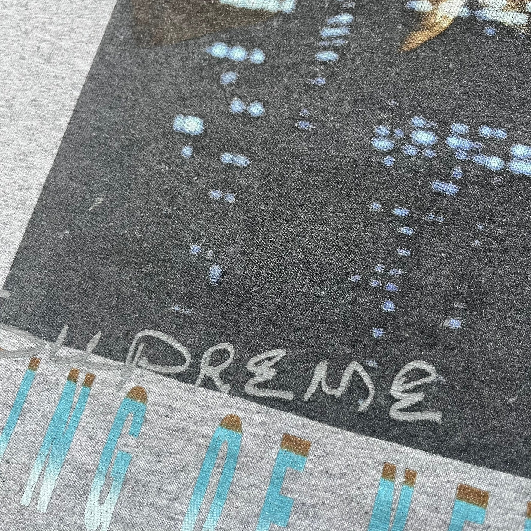 Supreme King of New York SS19 Gray Tee is a heather gray T-shirt released on February 21, 2019, as part of the SS19 collection. The front features an edited King of New York movie cover graphic with Christopher Walken’s name removed and replaced by handwritten-style “Supreme” and “C. Walken” text. The print is based on the original film cover artwork.