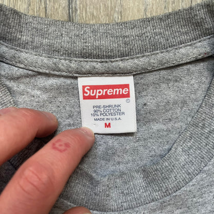 Supreme King of New York SS19 Gray Tee is a heather gray T-shirt released on February 21, 2019, as part of the SS19 collection. The front features an edited King of New York movie cover graphic with Christopher Walken’s name removed and replaced by handwritten-style “Supreme” and “C. Walken” text. The print is based on the original film cover artwork.