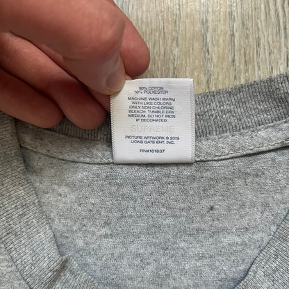 Supreme King of New York SS19 Gray Tee is a heather gray T-shirt released on February 21, 2019, as part of the SS19 collection. The front features an edited King of New York movie cover graphic with Christopher Walken’s name removed and replaced by handwritten-style “Supreme” and “C. Walken” text. The print is based on the original film cover artwork.