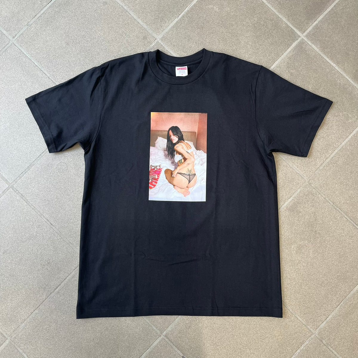 Product description:
Supreme Larry Clark FW26 Black Neighbor Tee is a black, 100% cotton T-shirt featuring a front photograph by Larry Clark of Gabbriette (Gabriella Leigh Bechtel) seated in underwear on a bed beside a red Supreme skateboard. The back displays hand-drawn-style white text reading "Love Thy Neighbor, Larry Clark 2026, Supreme."