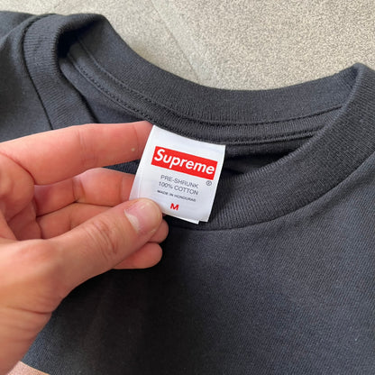Supreme Larry Clark FW26 Black Neighbor Tee is a black, 100% cotton T-shirt featuring a front photograph by Larry Clark of Gabbriette (Gabriella Leigh Bechtel) seated in underwear on a bed beside a red Supreme skateboard. The back displays hand-drawn-style white text reading "Love Thy Neighbor, Larry Clark 2026, Supreme."