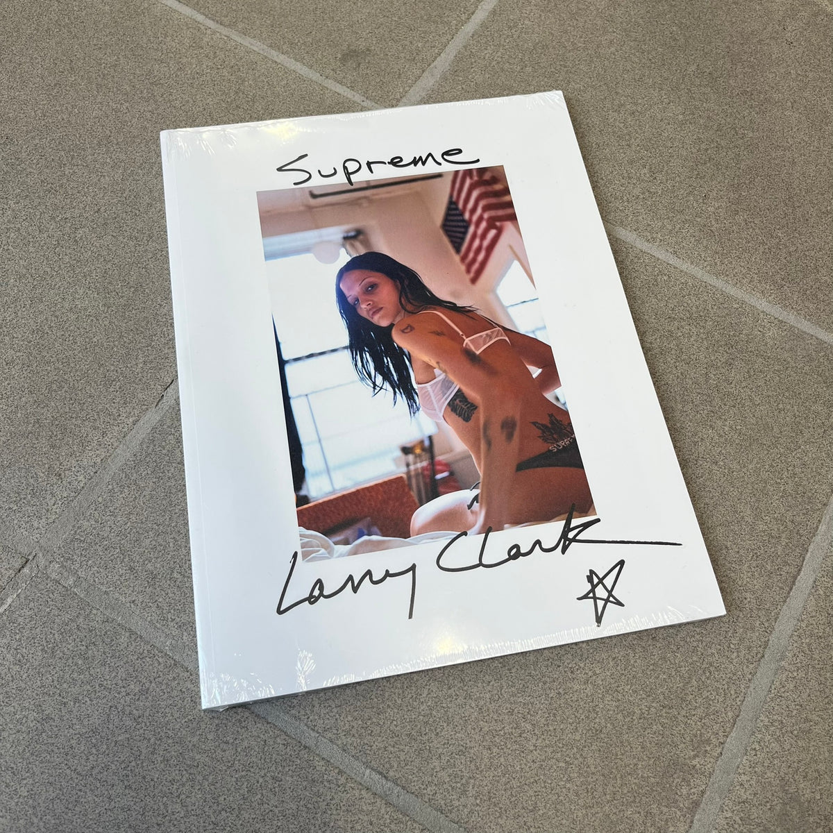 Supreme Larry Clark FW26 Multicolor Zine is a 48-page white zine with original photography by Larry Clark, including a poster insert. The cover features Gabbriette (Gabriella Leigh Bechtel) styled in Supreme underwear against a USA flag backdrop, with hand-drawn "Supreme" text scribbled at the top and "Larry Clark" with a star at the bottom, while the back displays a small Supreme logo.