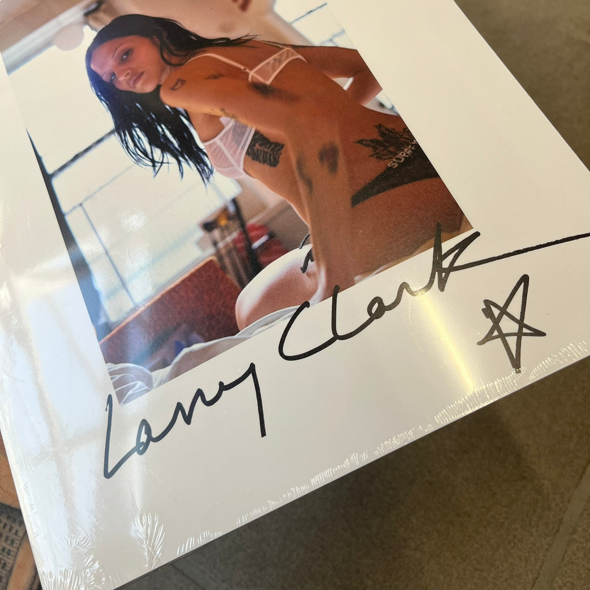 Supreme Larry Clark FW26 Multicolor Zine is a 48-page white zine with original photography by Larry Clark, including a poster insert. The cover features Gabbriette (Gabriella Leigh Bechtel) styled in Supreme underwear against a USA flag backdrop, with hand-drawn "Supreme" text scribbled at the top and "Larry Clark" with a star at the bottom, while the back displays a small Supreme logo.