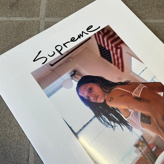 Supreme Larry Clark FW26 Multicolor Zine is a 48-page white zine with original photography by Larry Clark, including a poster insert. The cover features Gabbriette (Gabriella Leigh Bechtel) styled in Supreme underwear against a USA flag backdrop, with hand-drawn "Supreme" text scribbled at the top and "Larry Clark" with a star at the bottom, while the back displays a small Supreme logo.