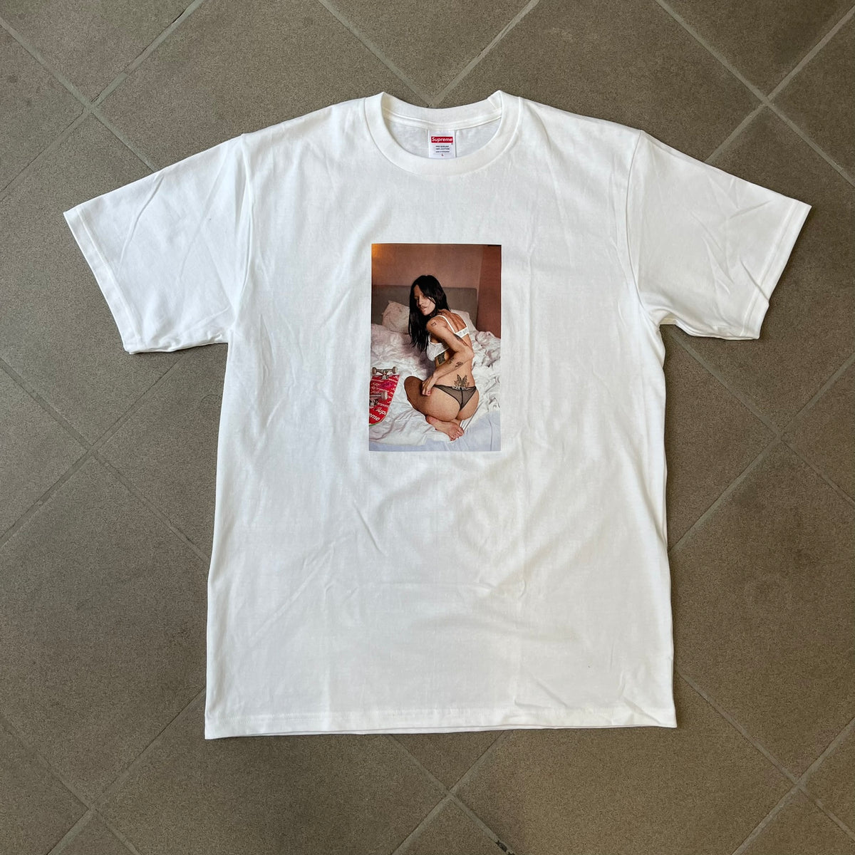 Supreme Larry Clark FW26 White Neighbor Tee is a white, 100% cotton T-shirt featuring a front photograph by Larry Clark of Gabbriette (Gabriella Leigh Bechtel) seated in underwear on a bed beside a red Supreme skateboard. The back displays hand-drawn-style black text reading "Love Thy Neighbor, Larry Clark 2026, Supreme."