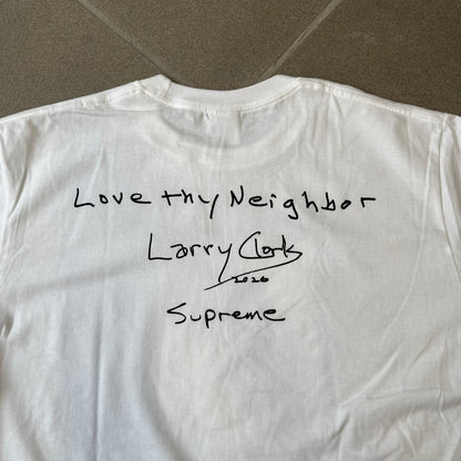 Supreme Larry Clark FW26 White Neighbor Tee is a white, 100% cotton T-shirt featuring a front photograph by Larry Clark of Gabbriette (Gabriella Leigh Bechtel) seated in underwear on a bed beside a red Supreme skateboard. The back displays hand-drawn-style black text reading "Love Thy Neighbor, Larry Clark 2026, Supreme."
