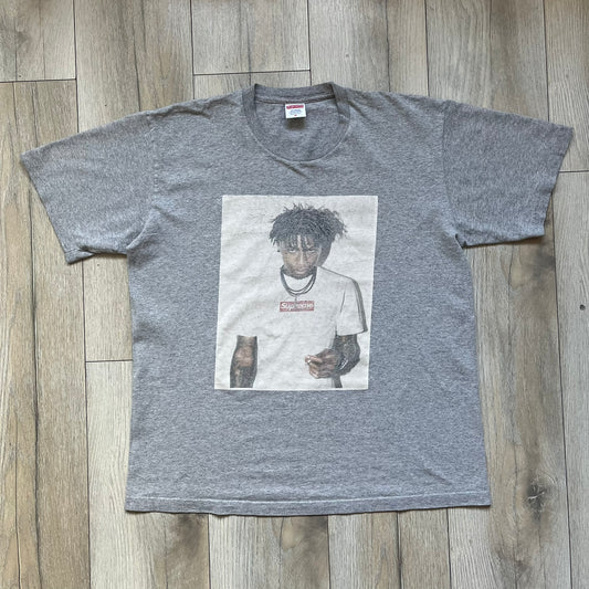 Supreme NBA Youngboy FW23 Gray Tee is a gray crew-neck T-shirt made in the USA from 100% cotton. The front features a multicolor graphic print of NBA Youngboy wearing the Supreme Box Logo tee. The shirt has short sleeves and a regular fit.