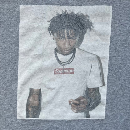 Supreme NBA Youngboy FW23 Gray Tee is a gray crew-neck T-shirt made in the USA from 100% cotton. The front features a multicolor graphic print of NBA Youngboy wearing the Supreme Box Logo tee. The shirt has short sleeves and a regular fit.