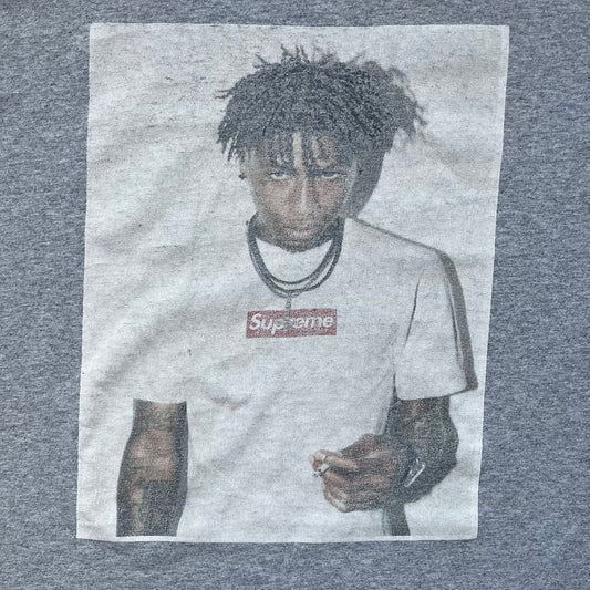 Supreme NBA Youngboy FW23 Gray Tee is a gray crew-neck T-shirt made in the USA from 100% cotton. The front features a multicolor graphic print of NBA Youngboy wearing the Supreme Box Logo tee. The shirt has short sleeves and a regular fit.