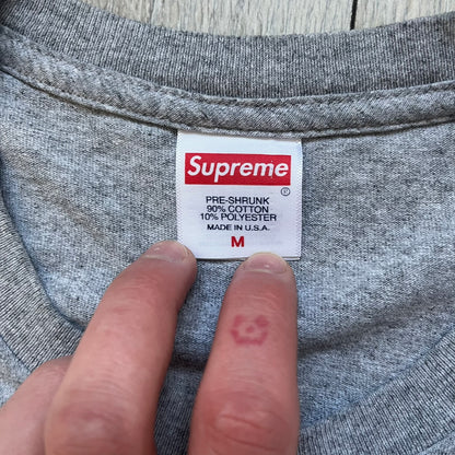 Supreme NBA Youngboy FW23 Gray Tee is a gray crew-neck T-shirt made in the USA from 100% cotton. The front features a multicolor graphic print of NBA Youngboy wearing the Supreme Box Logo tee. The shirt has short sleeves and a regular fit.