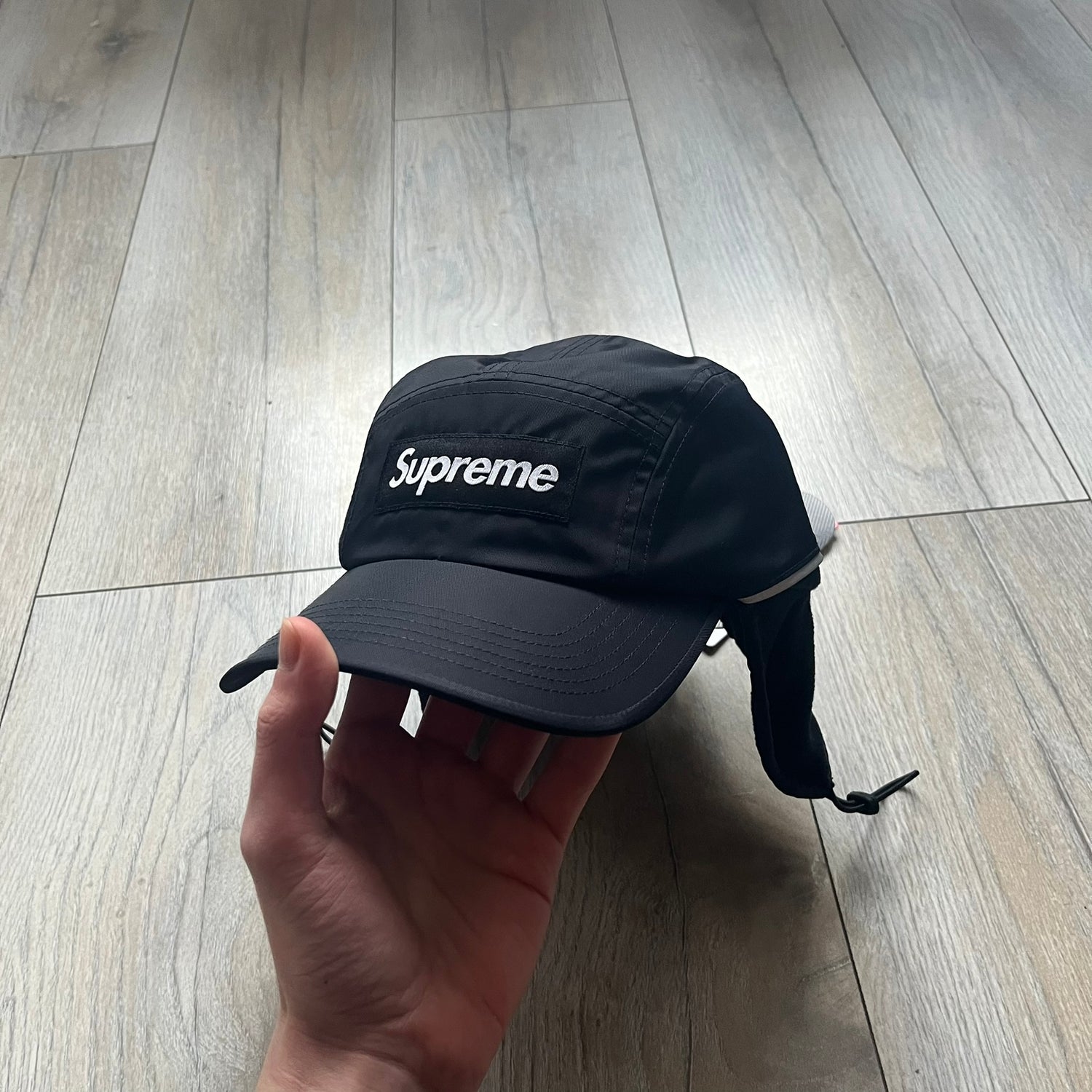 Supreme FW25 camp cap constructed from water-resistant Cordura® Advanced Fabrics nylon with a nylon and polyester shell. Lined with Polartec® 200 fleece for insulation, featuring packable earflaps secured with an interior elastic shockcord. Details include a webbing strap closure, 3M reflective piping, embroidered logos on the sides, and a classic Supreme box logo at the front. Finished in black.