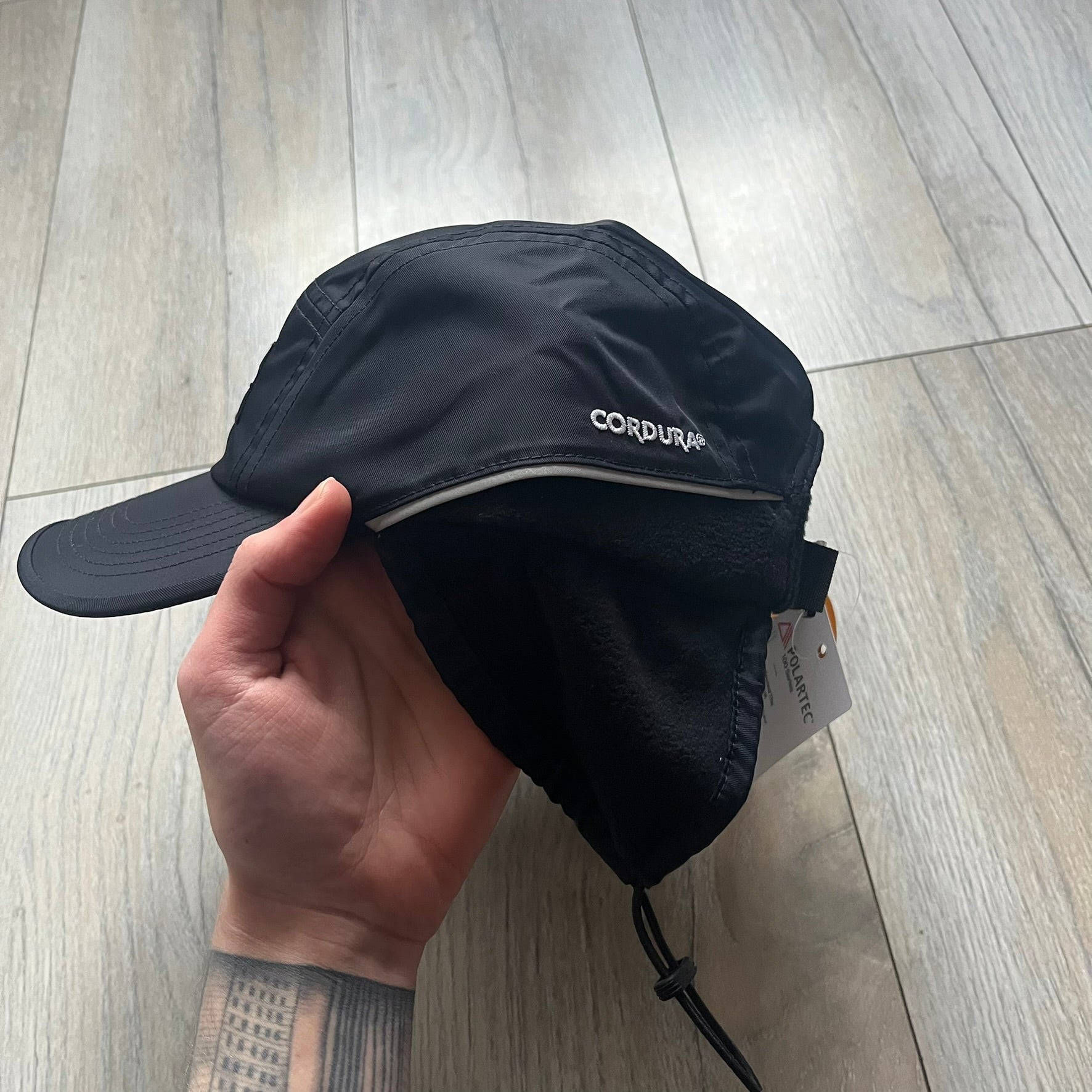 Supreme FW25 camp cap constructed from water-resistant Cordura® Advanced Fabrics nylon with a nylon and polyester shell. Lined with Polartec® 200 fleece for insulation, featuring packable earflaps secured with an interior elastic shockcord. Details include a webbing strap closure, 3M reflective piping, embroidered logos on the sides, and a classic Supreme box logo at the front. Finished in black.