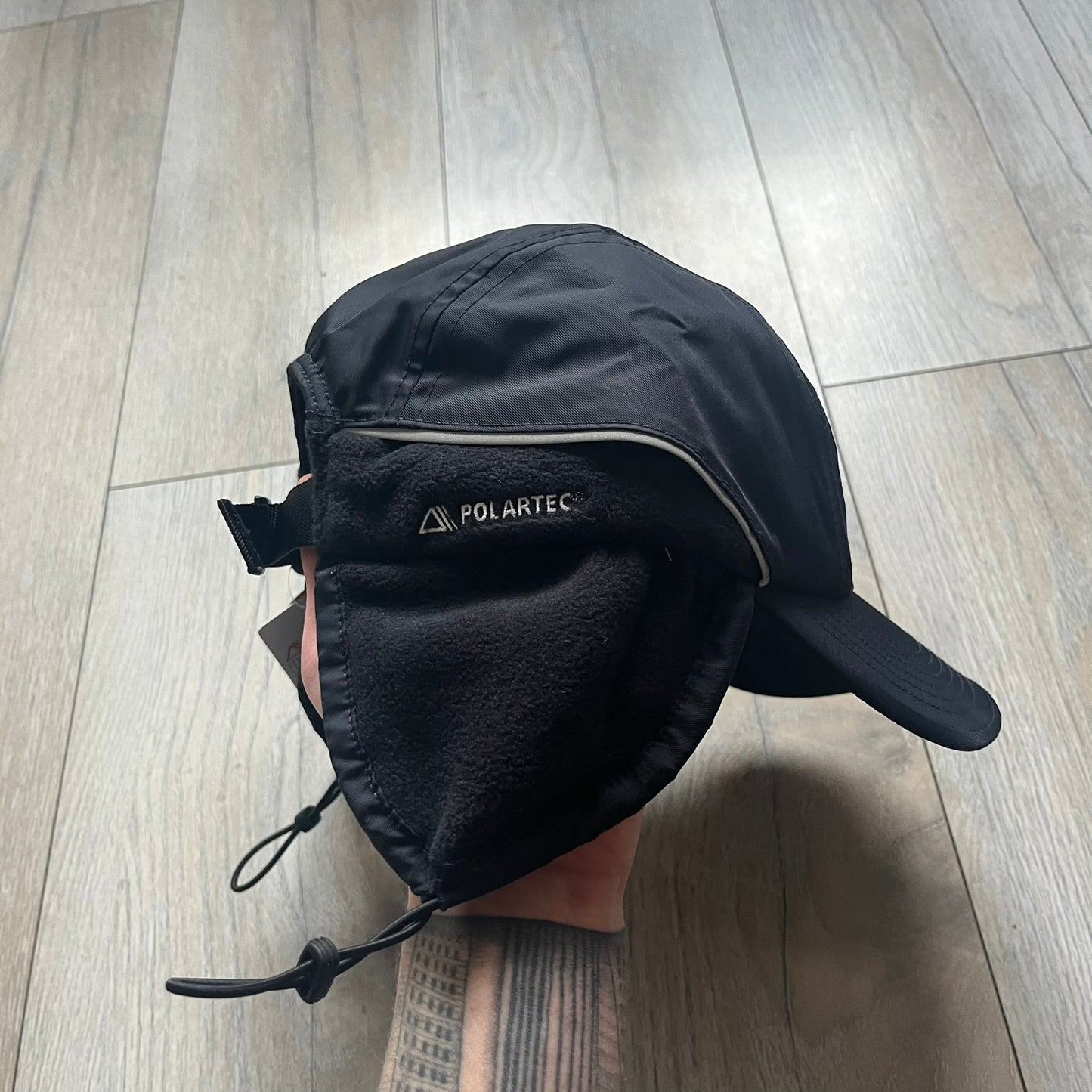 Supreme FW25 camp cap constructed from water-resistant Cordura® Advanced Fabrics nylon with a nylon and polyester shell. Lined with Polartec® 200 fleece for insulation, featuring packable earflaps secured with an interior elastic shockcord. Details include a webbing strap closure, 3M reflective piping, embroidered logos on the sides, and a classic Supreme box logo at the front. Finished in black.