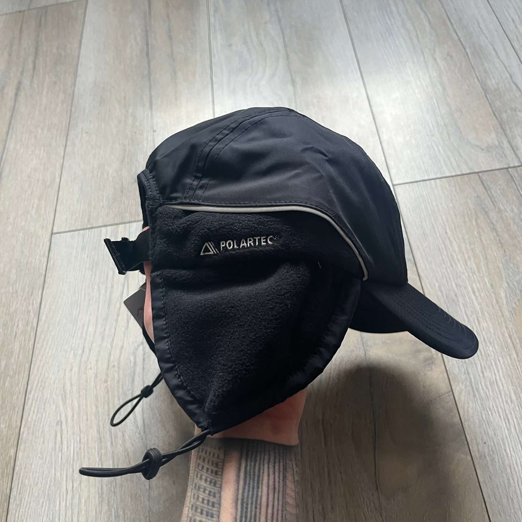 Supreme FW25 camp cap constructed from water-resistant Cordura® Advanced Fabrics nylon with a nylon and polyester shell. Lined with Polartec® 200 fleece for insulation, featuring packable earflaps secured with an interior elastic shockcord. Details include a webbing strap closure, 3M reflective piping, embroidered logos on the sides, and a classic Supreme box logo at the front. Finished in black.