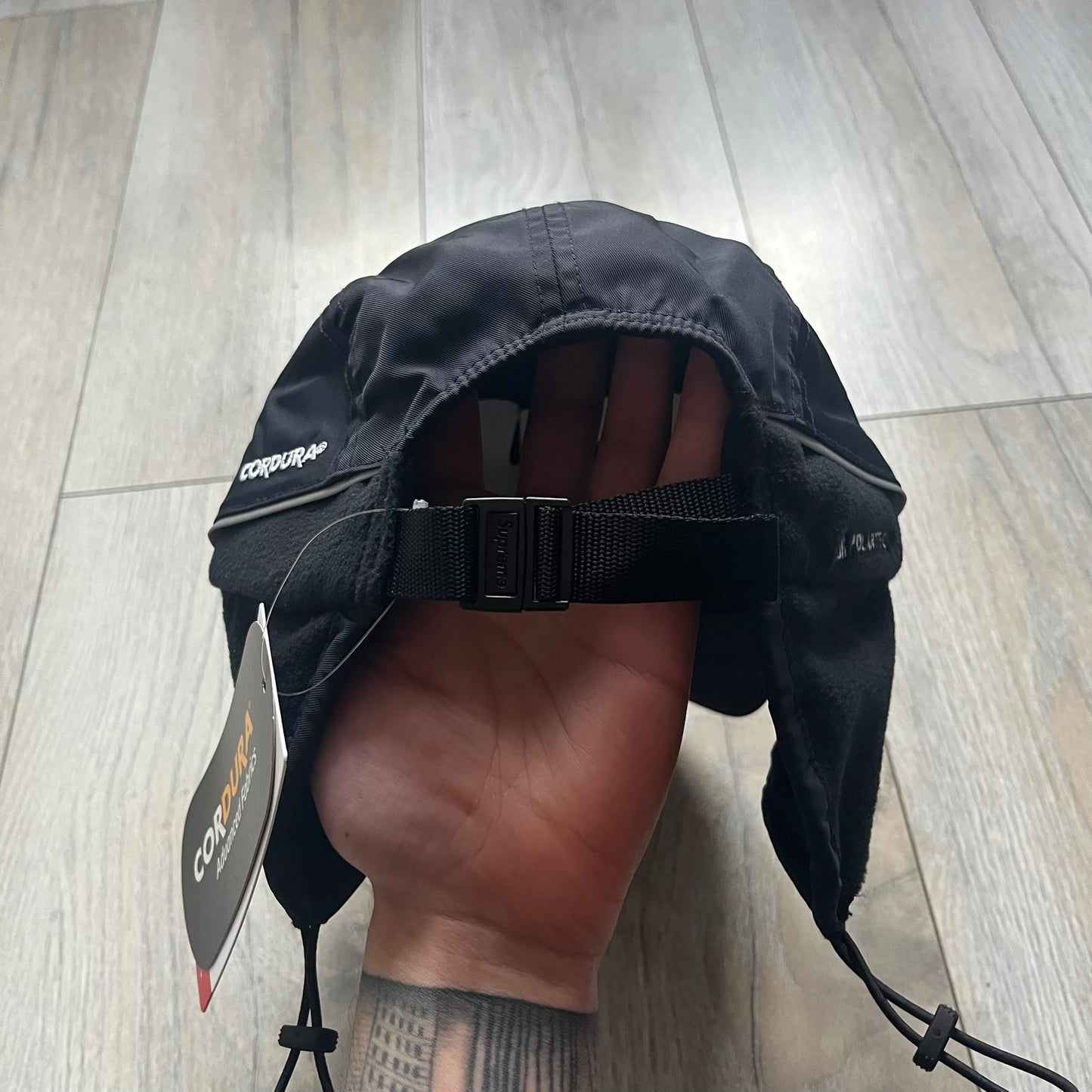 Supreme FW25 camp cap constructed from water-resistant Cordura® Advanced Fabrics nylon with a nylon and polyester shell. Lined with Polartec® 200 fleece for insulation, featuring packable earflaps secured with an interior elastic shockcord. Details include a webbing strap closure, 3M reflective piping, embroidered logos on the sides, and a classic Supreme box logo at the front. Finished in black.