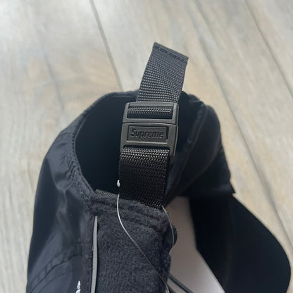 Supreme FW25 camp cap constructed from water-resistant Cordura® Advanced Fabrics nylon with a nylon and polyester shell. Lined with Polartec® 200 fleece for insulation, featuring packable earflaps secured with an interior elastic shockcord. Details include a webbing strap closure, 3M reflective piping, embroidered logos on the sides, and a classic Supreme box logo at the front. Finished in black.