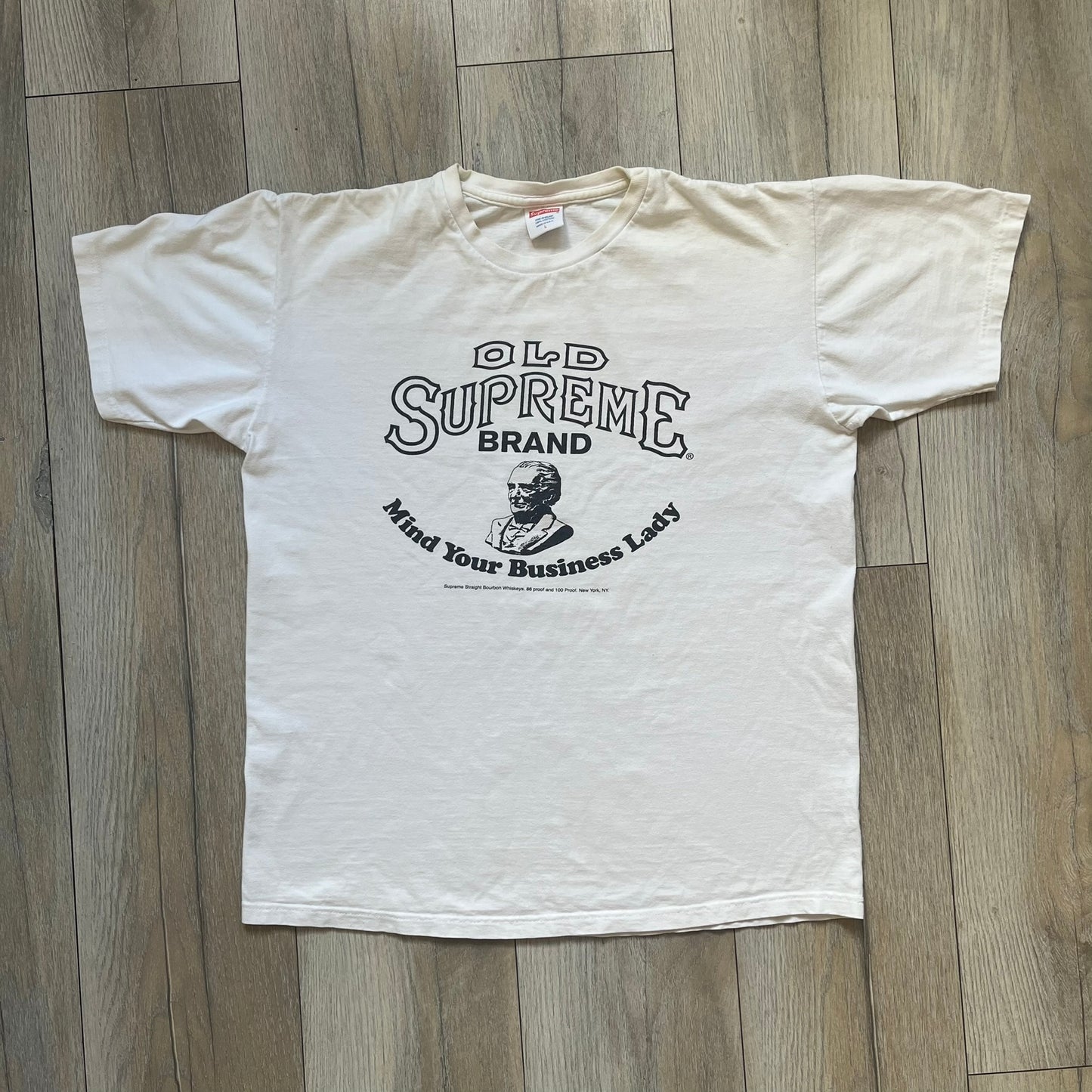 Supreme SS11 Mind Your Business White Tee is a white short-sleeve T-shirt from the Spring/Summer 2011 collection, made in the USA. The front features a graphic inspired by a whiskey label, including an old Supreme logo, a bust of an older man, a small “Mind Your Business” figure, and text referencing Supreme Bourbon Whiskeys with a New York address. This design is part of Supreme’s second delivery of SS11 T-shirts.