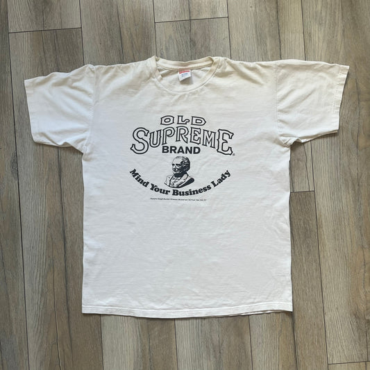 Supreme SS11 Mind Your Business White Tee is a white short-sleeve T-shirt from the Spring/Summer 2011 collection, made in the USA. The front features a graphic inspired by a whiskey label, including an old Supreme logo, a bust of an older man, a small “Mind Your Business” figure, and text referencing Supreme Bourbon Whiskeys with a New York address. This design is part of Supreme’s second delivery of SS11 T-shirts.