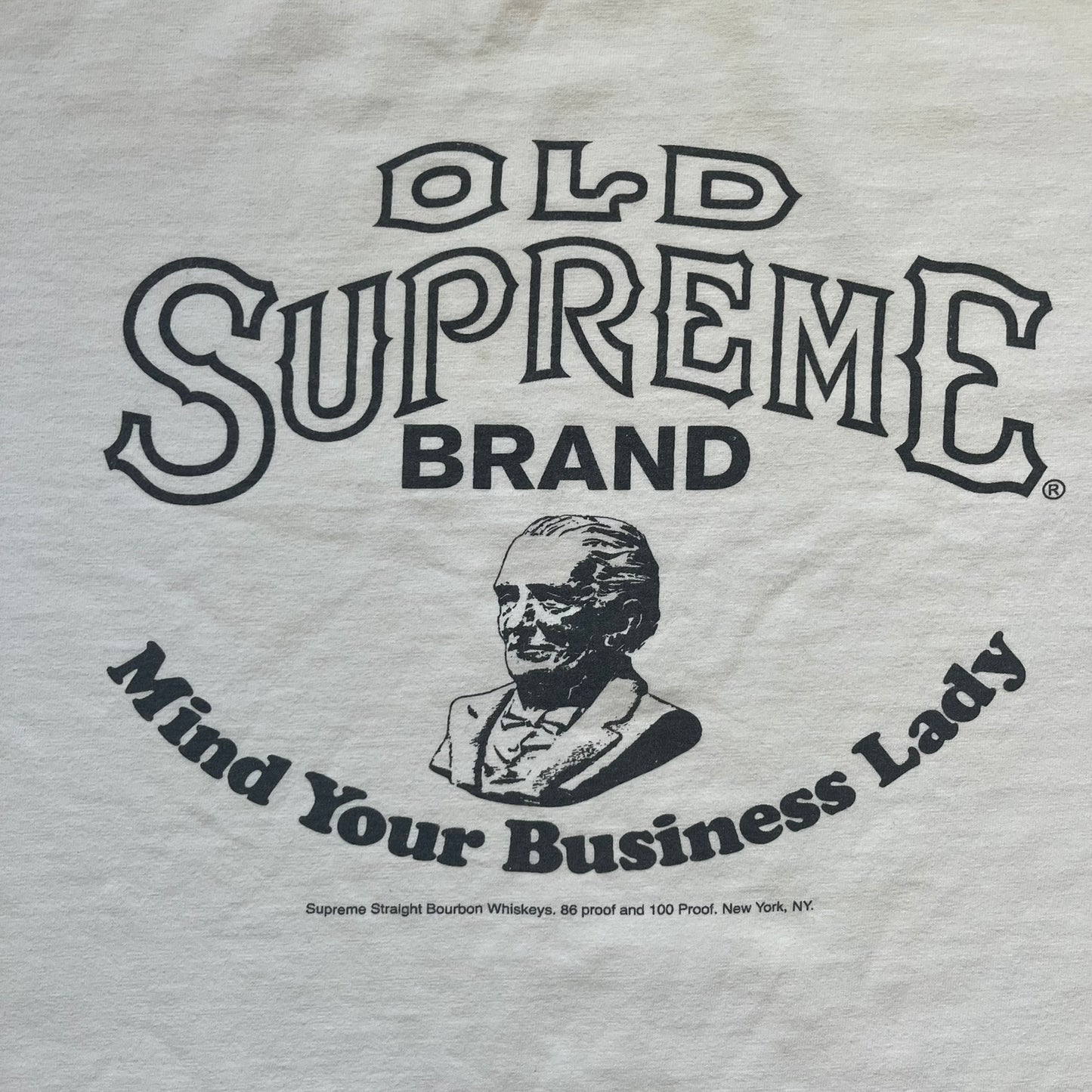 Supreme SS11 Mind Your Business White Tee is a white short-sleeve T-shirt from the Spring/Summer 2011 collection, made in the USA. The front features a graphic inspired by a whiskey label, including an old Supreme logo, a bust of an older man, a small “Mind Your Business” figure, and text referencing Supreme Bourbon Whiskeys with a New York address. This design is part of Supreme’s second delivery of SS11 T-shirts.