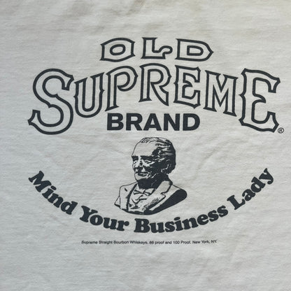 Supreme SS11 Mind Your Business White Tee is a white short-sleeve T-shirt from the Spring/Summer 2011 collection, made in the USA. The front features a graphic inspired by a whiskey label, including an old Supreme logo, a bust of an older man, a small “Mind Your Business” figure, and text referencing Supreme Bourbon Whiskeys with a New York address. This design is part of Supreme’s second delivery of SS11 T-shirts.