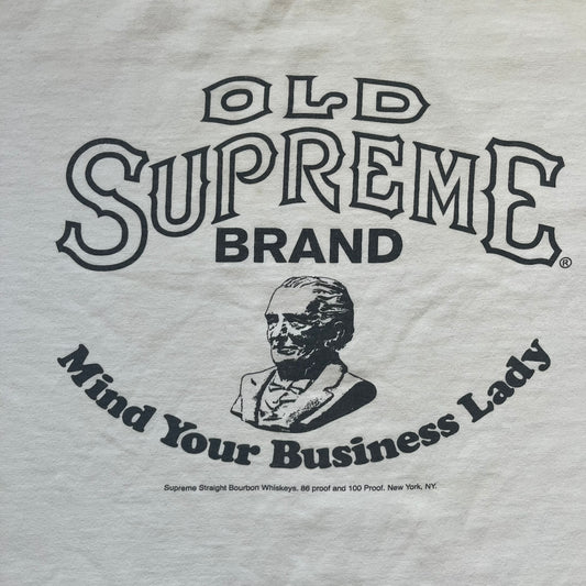 Supreme SS11 Mind Your Business White Tee is a white short-sleeve T-shirt from the Spring/Summer 2011 collection, made in the USA. The front features a graphic inspired by a whiskey label, including an old Supreme logo, a bust of an older man, a small “Mind Your Business” figure, and text referencing Supreme Bourbon Whiskeys with a New York address. This design is part of Supreme’s second delivery of SS11 T-shirts.