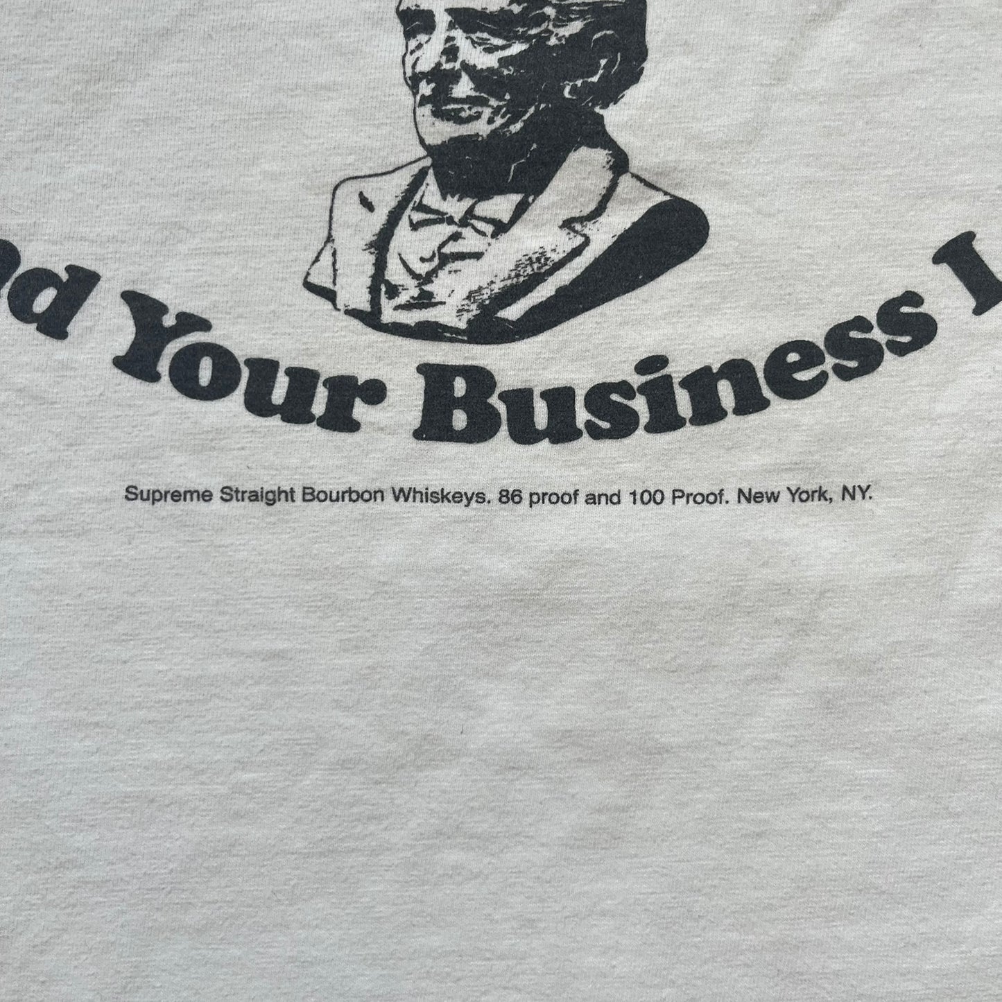 Supreme SS11 Mind Your Business White Tee is a white short-sleeve T-shirt from the Spring/Summer 2011 collection, made in the USA. The front features a graphic inspired by a whiskey label, including an old Supreme logo, a bust of an older man, a small “Mind Your Business” figure, and text referencing Supreme Bourbon Whiskeys with a New York address. This design is part of Supreme’s second delivery of SS11 T-shirts.