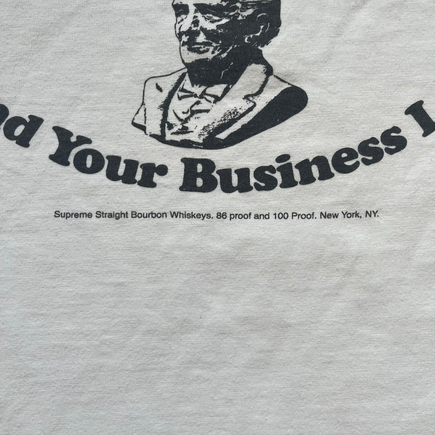 Supreme SS11 Mind Your Business White Tee is a white short-sleeve T-shirt from the Spring/Summer 2011 collection, made in the USA. The front features a graphic inspired by a whiskey label, including an old Supreme logo, a bust of an older man, a small “Mind Your Business” figure, and text referencing Supreme Bourbon Whiskeys with a New York address. This design is part of Supreme’s second delivery of SS11 T-shirts.
