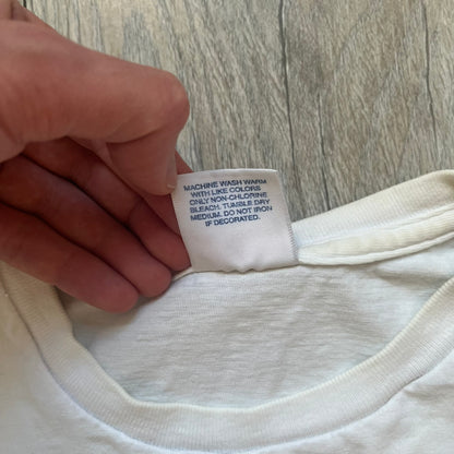 Supreme SS11 Mind Your Business White Tee is a white short-sleeve T-shirt from the Spring/Summer 2011 collection, made in the USA. The front features a graphic inspired by a whiskey label, including an old Supreme logo, a bust of an older man, a small “Mind Your Business” figure, and text referencing Supreme Bourbon Whiskeys with a New York address. This design is part of Supreme’s second delivery of SS11 T-shirts.