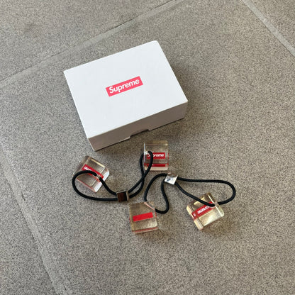 Supreme SS18 Hair Bobbles (Set of 2) features clear acrylic dice-shaped pieces with small red Supreme Box Logo details inside, attached to black elastic bands. The set includes two hair bobbles and comes in the original white Supreme box. Released on May 17, 2018.
