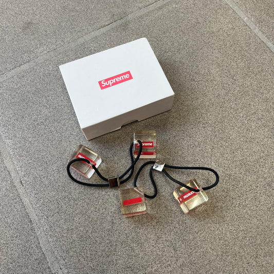 Supreme SS18 Hair Bobbles (Set of 2) features clear acrylic dice-shaped pieces with small red Supreme Box Logo details inside, attached to black elastic bands. The set includes two hair bobbles and comes in the original white Supreme box. Released on May 17, 2018.
