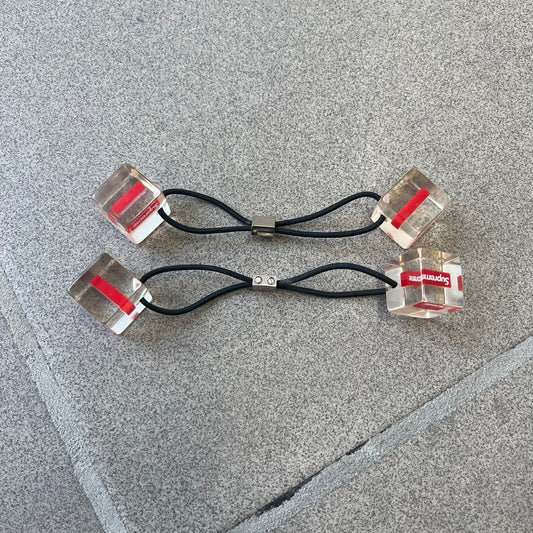 Supreme SS18 Hair Bobbles (Set of 2) features clear acrylic dice-shaped pieces with small red Supreme Box Logo details inside, attached to black elastic bands. The set includes two hair bobbles and comes in the original white Supreme box. Released on May 17, 2018.
