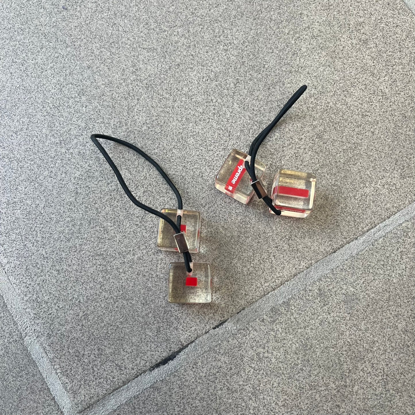 Supreme SS18 Hair Bobbles (Set of 2) features clear acrylic dice-shaped pieces with small red Supreme Box Logo details inside, attached to black elastic bands. The set includes two hair bobbles and comes in the original white Supreme box. Released on May 17, 2018.
