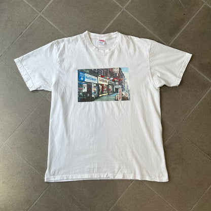 Supreme SS18 Hardware White Tee features a white 100% cotton T-shirt made in the USA. The front has a street scene with various storefronts, including a hardware store with a Supreme logo above it. A small Supreme box logo is printed on the upper back below the collar.

